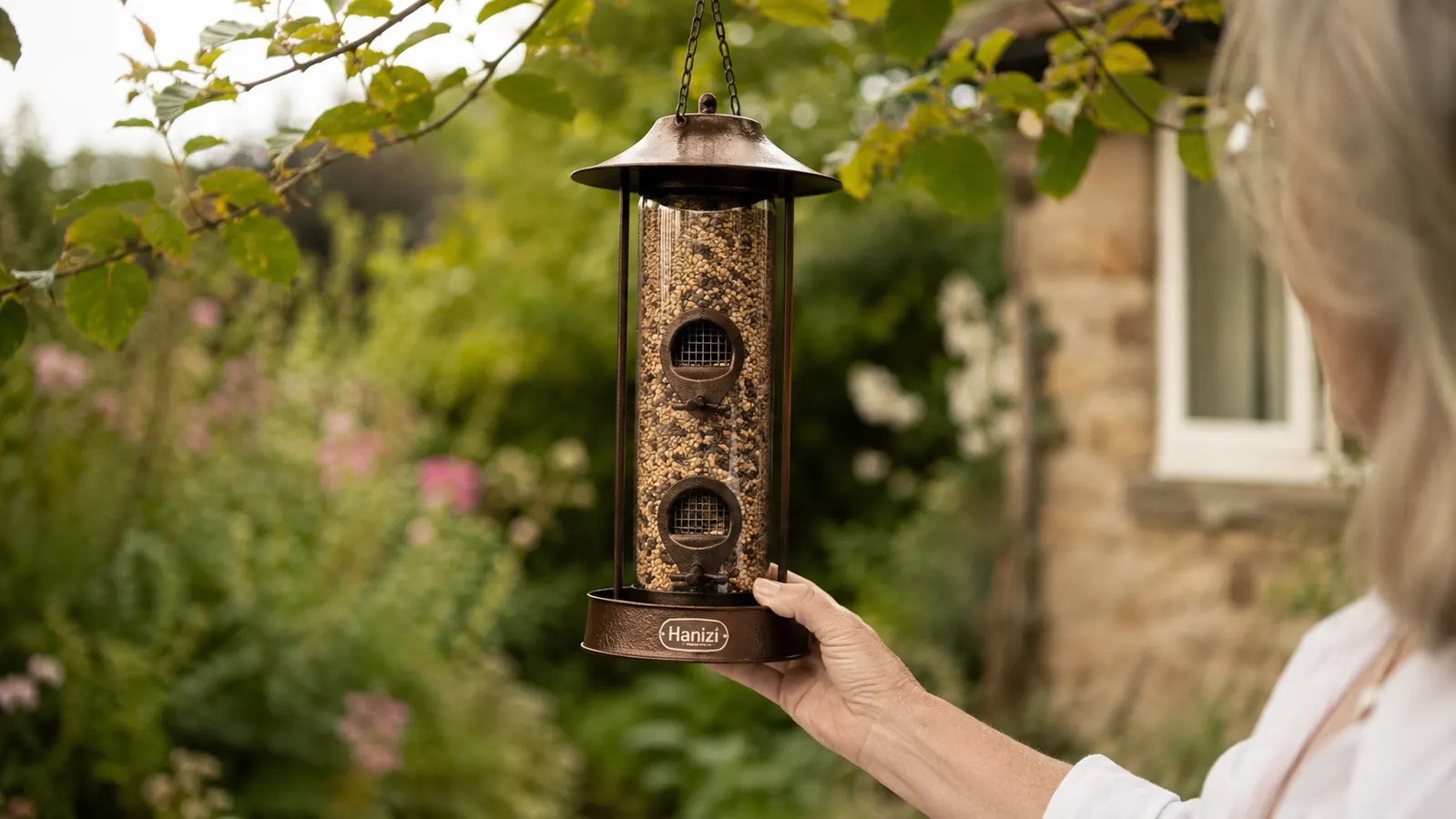 Hanizi brown hanging bird feeder filled with mixed seed, hand holding it up against a garden backdrop